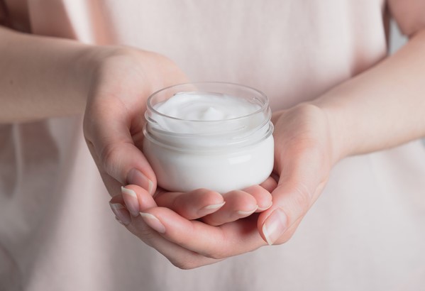 Womans hand holding a creamy lotion in a small glass makeup container