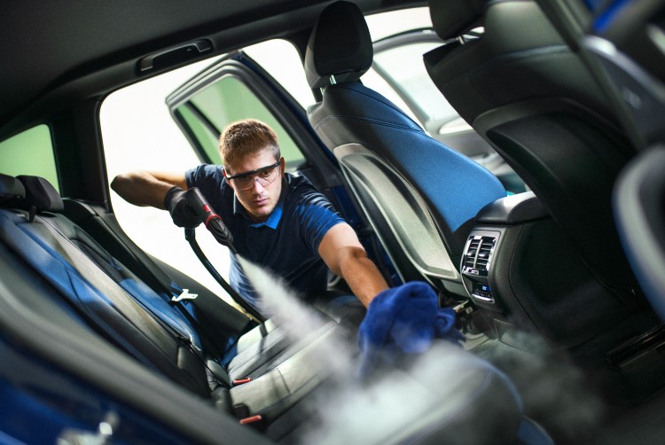 A car wash attendant leaning inside of a vehicle through the passenger door, spraying a cleaning solution on the leather upholstery. 