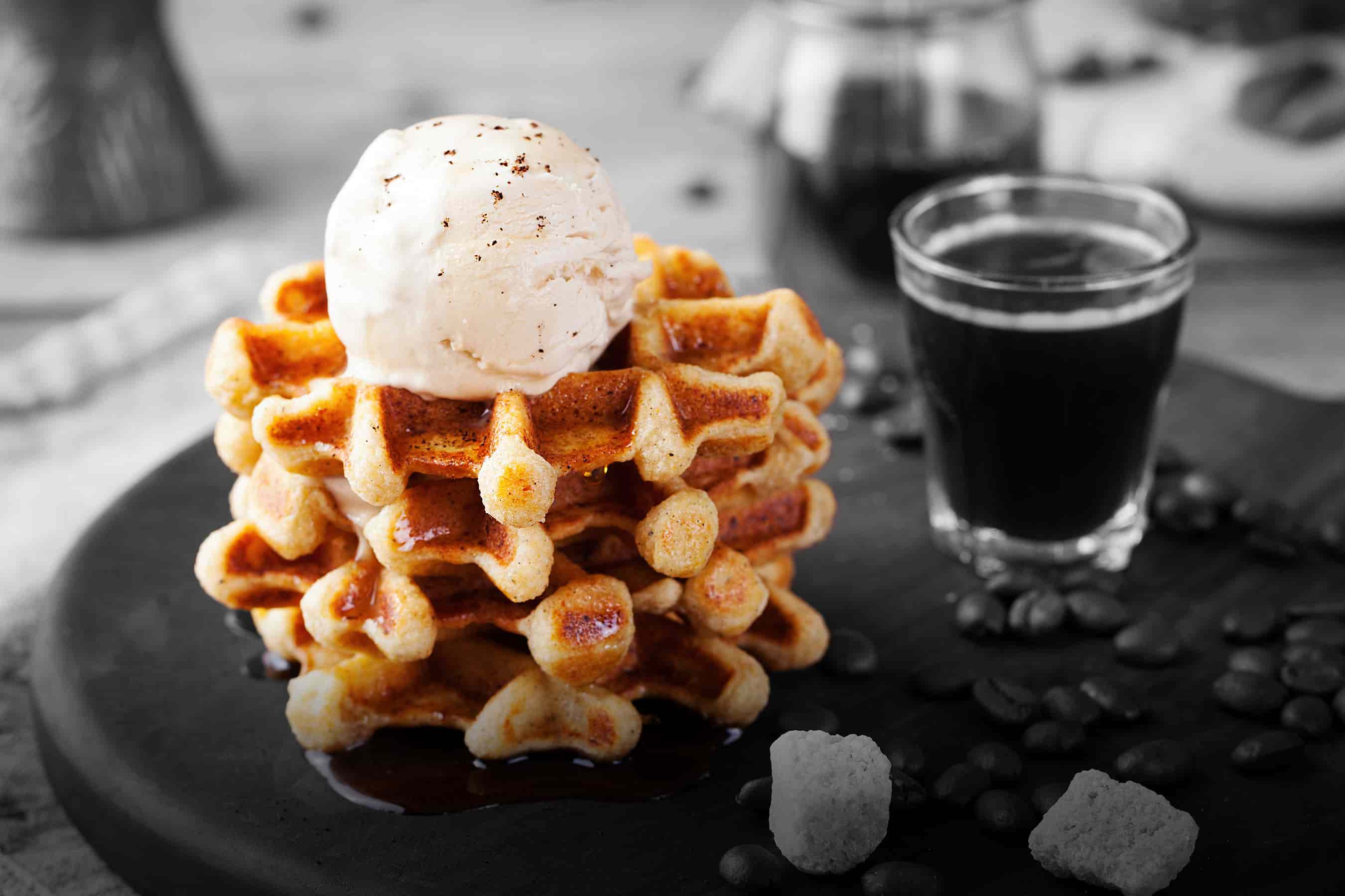 A stack of healthier ingredient waffles with a scoop of ice cream on top. A glass of espresso is displayed in the background.