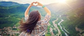 Woman on a hilltop making a heart shap with her hands over a majestic view