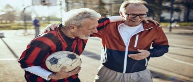 An older men laughing on a basketball court with a soccer ball in one hand and his arm around another laughing man