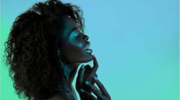 Side profile of a woman against a diffused green background touches her hand to her shimmering blue lip makeup