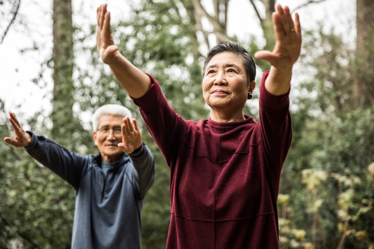 An older man and woman stretching in a forested area with arms outstretched.