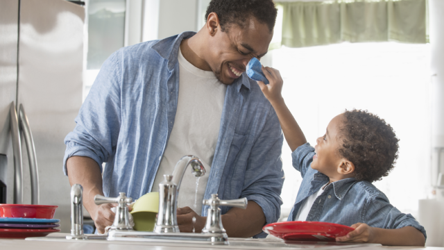 A  young son wipes water off of his laughing dads face while washing dishes at the kitchen sink
