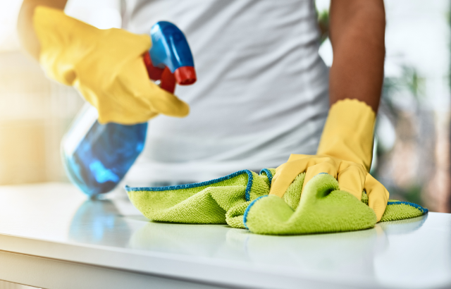 person wearing yellow cleaning gloves spraying solution on a surface while wiping it up with a microfiber cloth