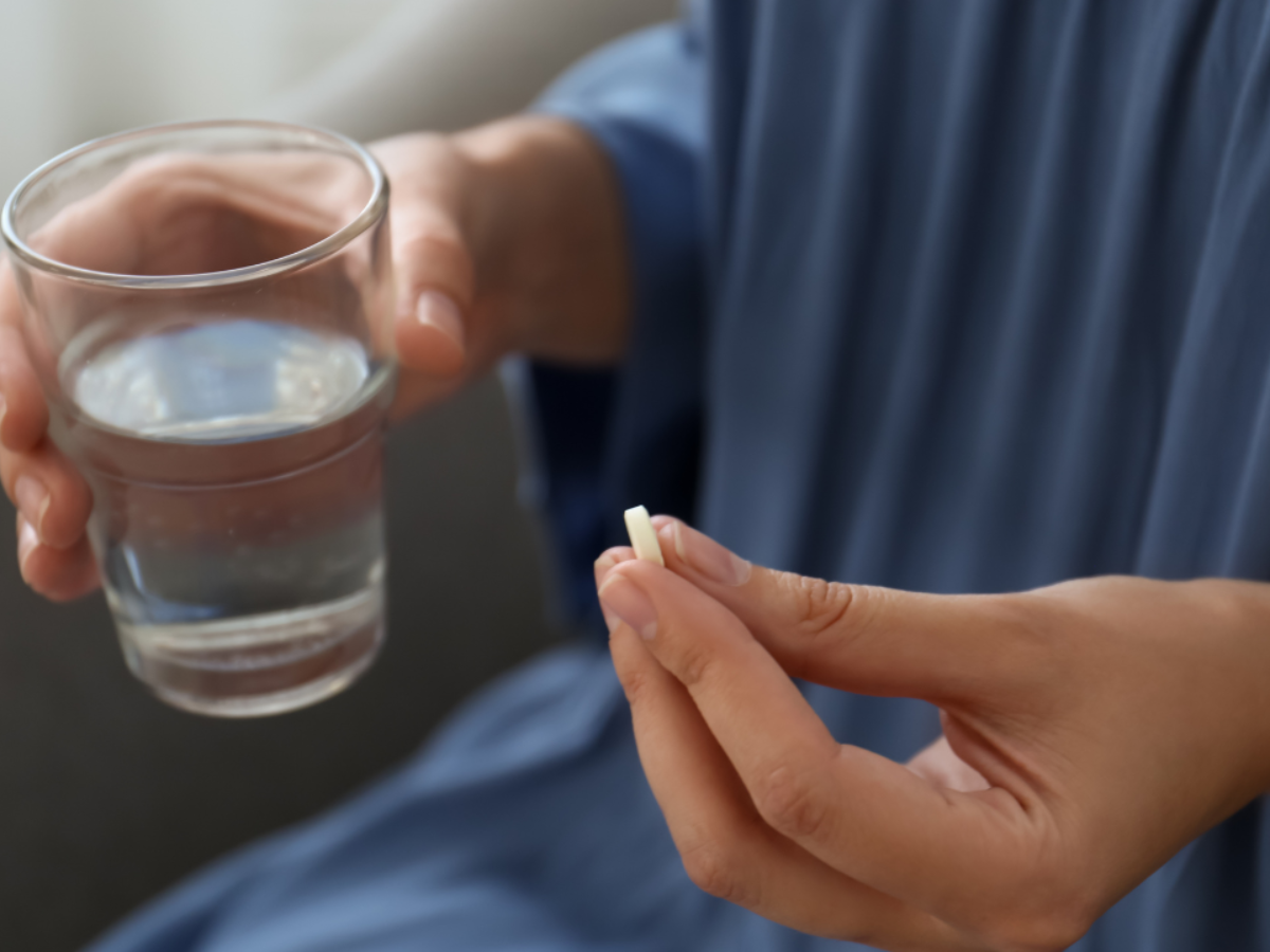 A pharmaceutical consumer's hands hold a prescription medication tablet in one hand and a glass of water in the other.