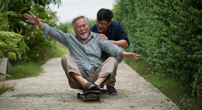 An older man happily sitting on a skateboard as a younger man gently pushes him down a sidewalk