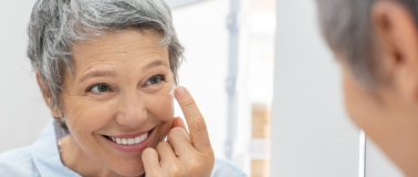 An older woman looks in a mirror as she applies skin care products