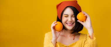 A woman wearing orange clothes stands in front of an orange background while laughing and holding an orange in each hand