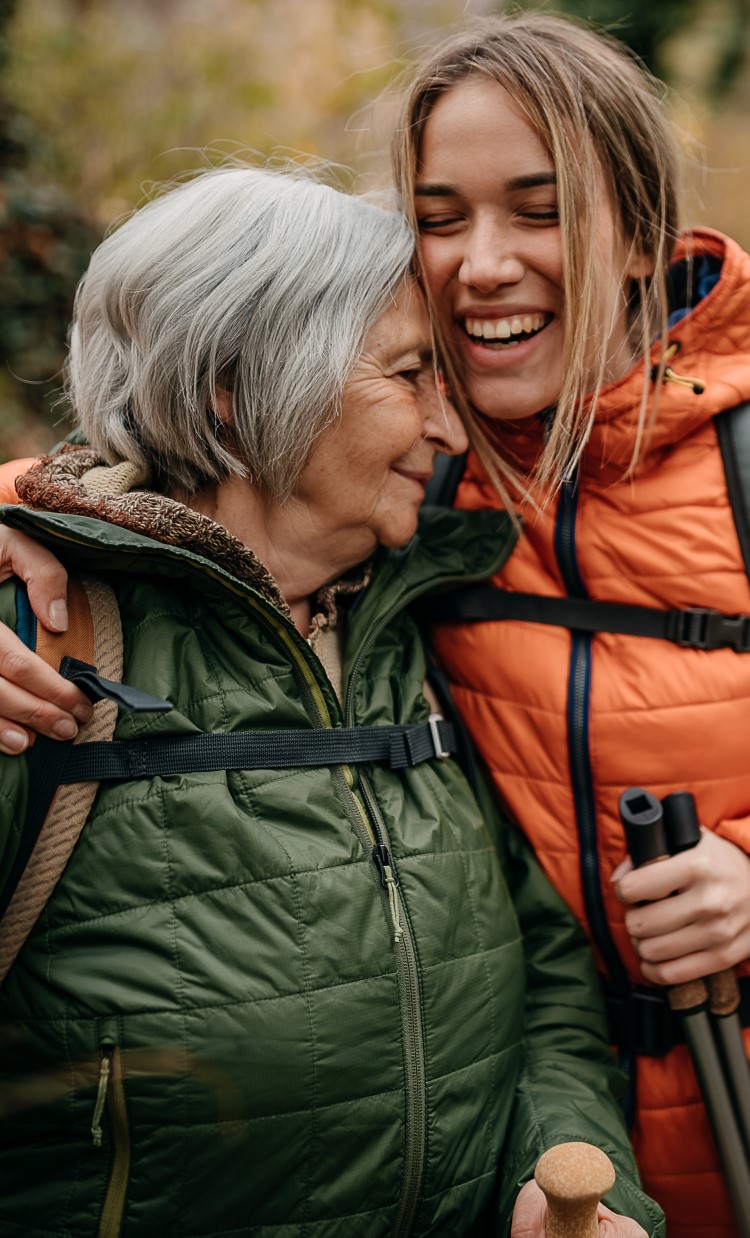 An older adult and younger adult outdoors with hiking gear hugging and laughing.
