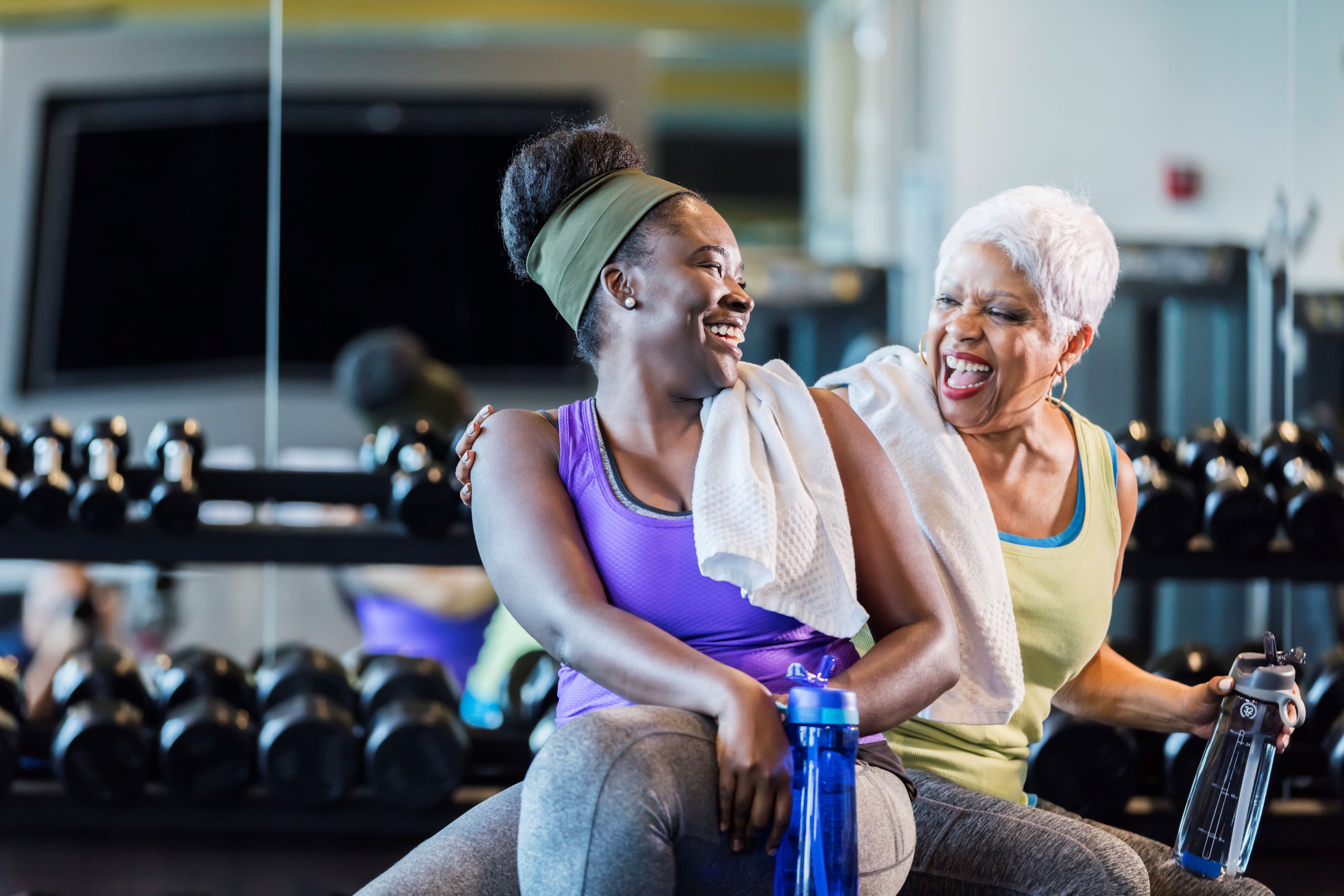 Two women share a laugh while working out together near free weights at the gym.